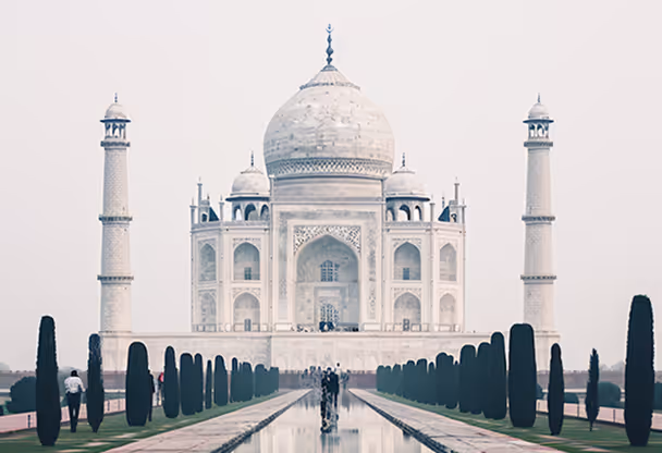 Front view of the Taj Mahal with its white marble dome, four minarets, and reflecting pool lined with tall, thin trees.