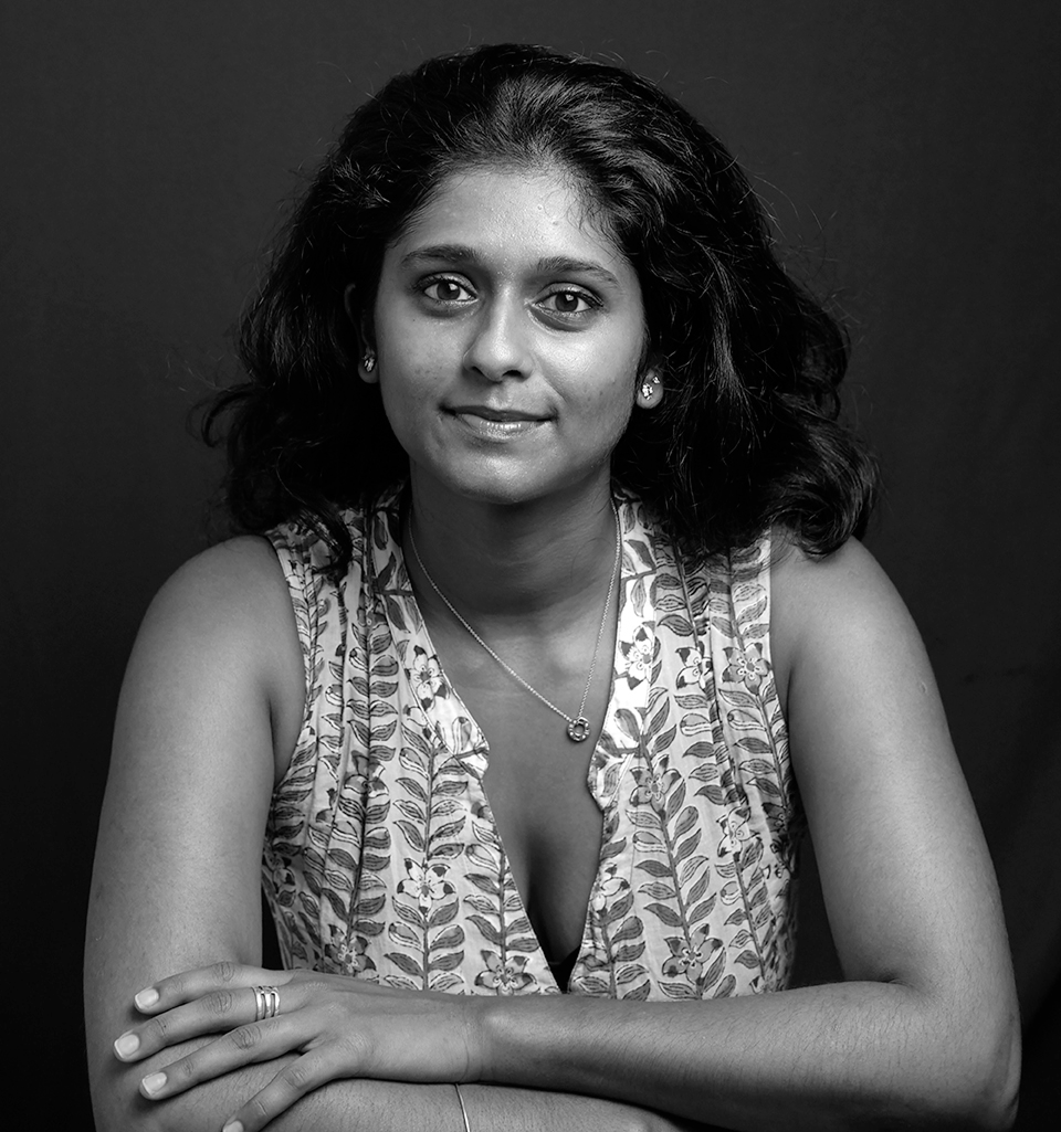 Woman with dark curly hair wearing a floral sleeveless top and a necklace, smiling calmly with arms crossed on a table.
