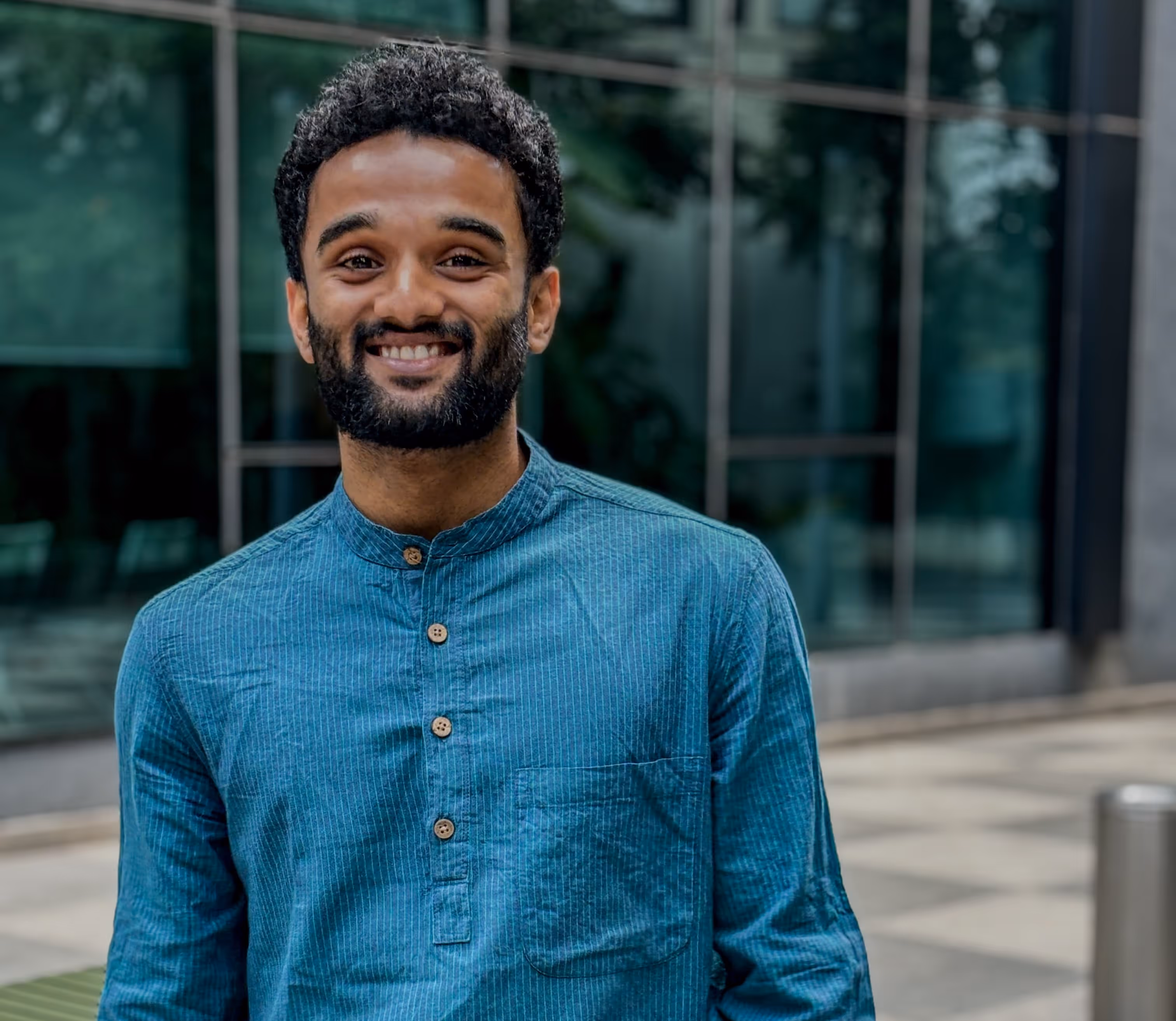 Smiling man with curly hair and beard wearing a blue shirt standing outdoors in front of a building with glass windows.